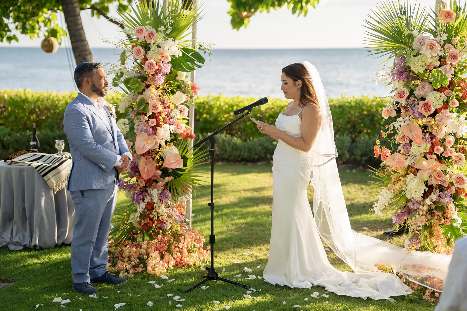 Becca reading her vows to Jake beneath the flower arches by the ocean