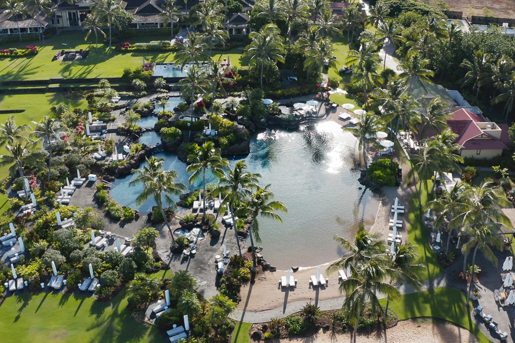 Aerial view of the Makai Pools at Kukui'ula — a sand-bottom lagoon, waterfalls and loungers ringed by coconut palms