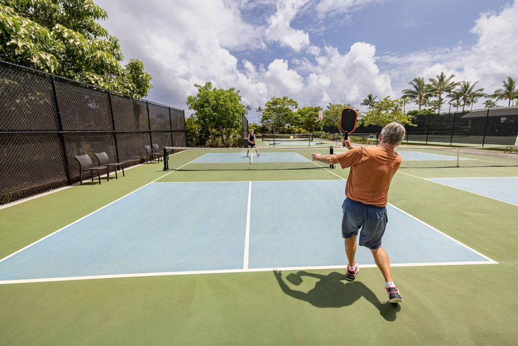 Two players volleying on one of the outdoor pickleball courts at Kukui'ula, palms and trade-wind clouds behind them