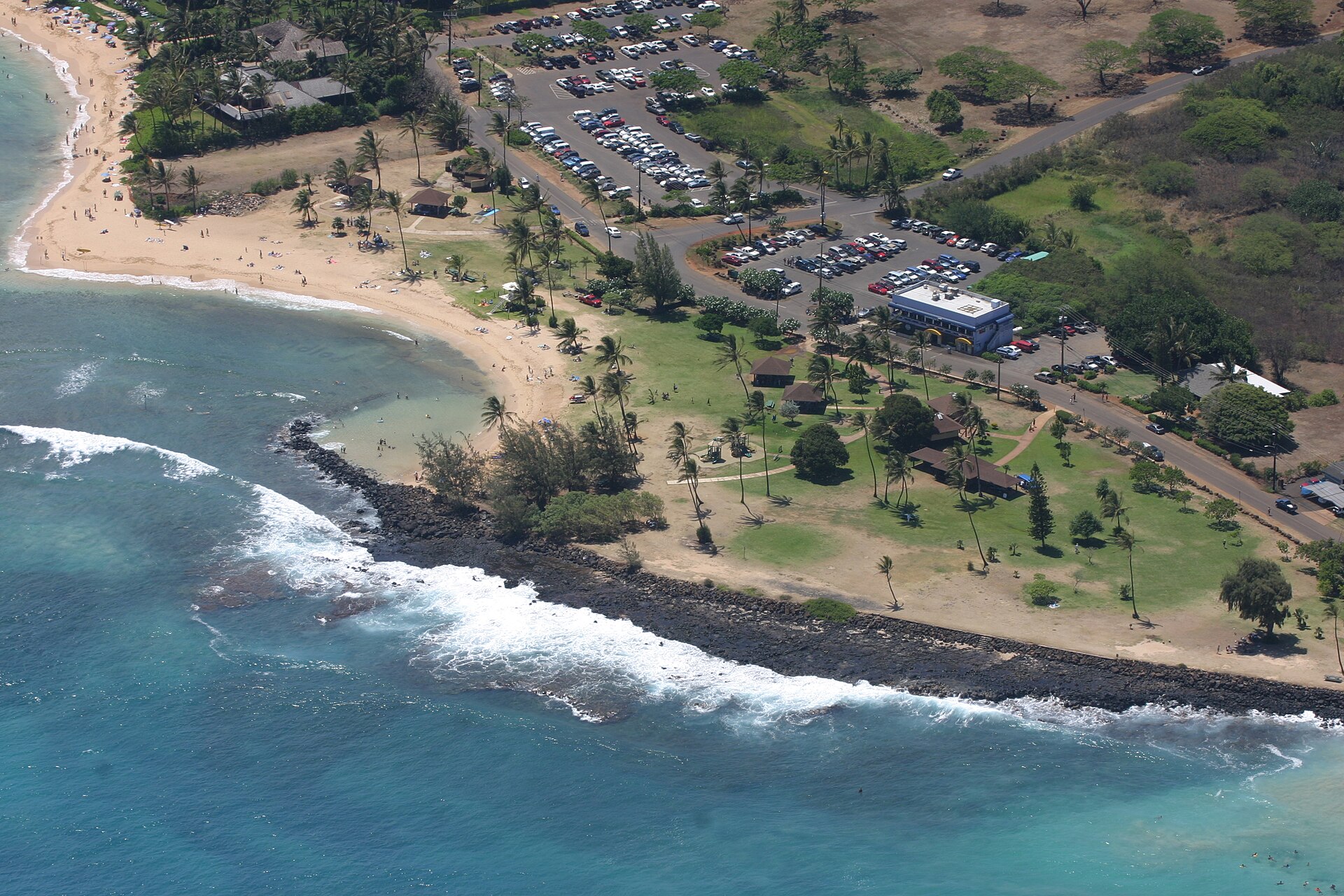 Aerial view of Pō'ipū Beach Park showing the twin crescents of sand meeting at a sandbar, with the parking lot and lawn behind